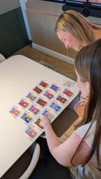 Two women leaning over a white meeting table in a modern room, smiling and pointing at a grid of colorful aura-style portrait prints spread across the table, one holding a drink.