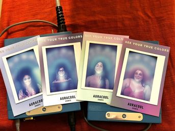 Four vibrant aura-photography instant prints of different people with colorful glowing halos, fanned across blue camera devices on bright red fabric — playful event photo keepsakes.