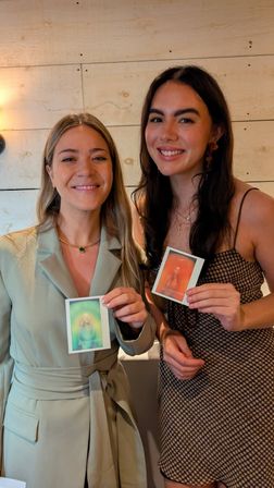 Two smiling women posing indoors against a light wood-paneled wall, each holding a colorful instant photo—one in a sage blazer, the other in a patterned dress; warm, casual studio vibe.