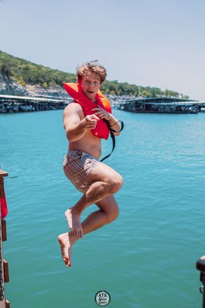 Man mid-jump off a lakeside dock into turquoise water, wearing a red life jacket and striped swim trunks, pointing at the camera with boat slips and tree-lined shore in the background.