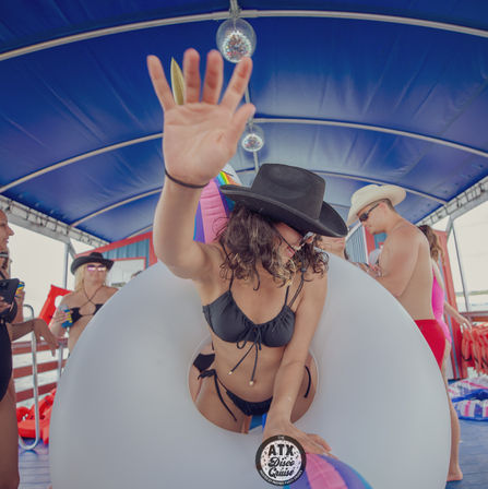 Party boat scene: person in black bikini and cowboy hat leaning through a giant white inner tube on a covered deck, disco balls overhead