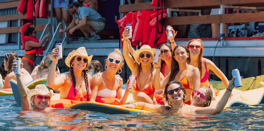 Group of friends in colorful bikinis and sun hats lounging on a large float near a wooden dock, smiling and raising canned drinks at a sunny summer waterfront party.