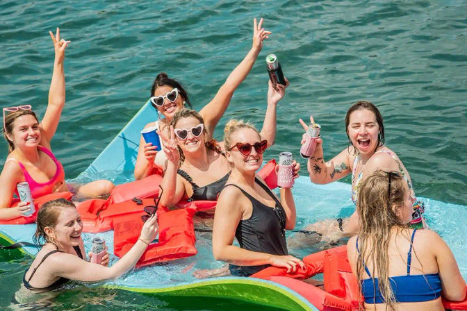 Group of friends partying on a colorful inflatable raft in a lake, wearing swimsuits and heart-shaped sunglasses, holding canned drinks and cheering on a sunny summer day
