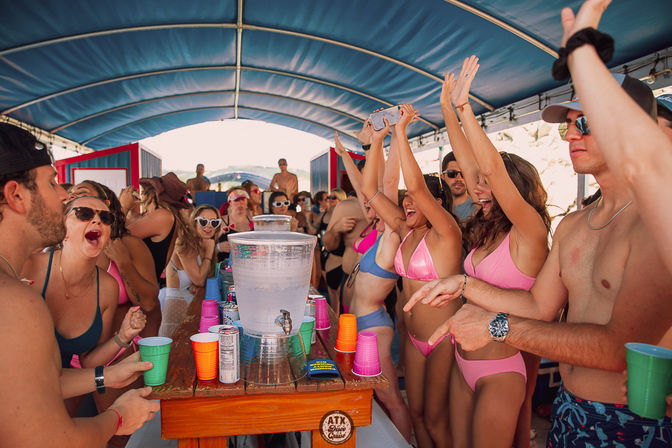 Young adults in swimsuits cheering around a drink table with colorful cups and a water dispenser on a covered party boat on a sunny summer day