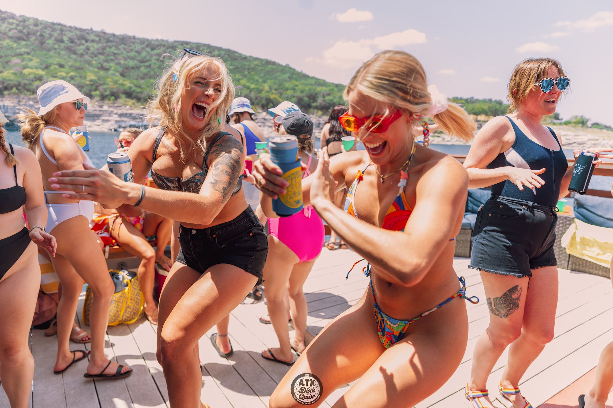 Women laughing and dancing on a sunny boat deck at a lakeside summer party, wearing swimsuits and holding canned drinks with water and tree-covered hills in the background.
