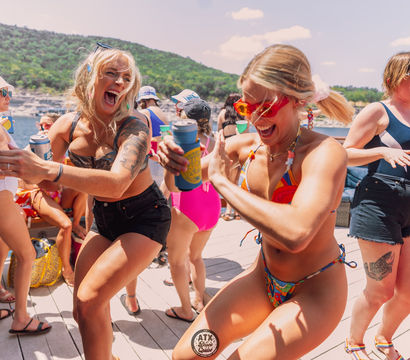 Women laughing and dancing on a sunny boat deck at a lakeside summer party, wearing swimsuits and holding canned drinks with water and tree-covered hills in the background.