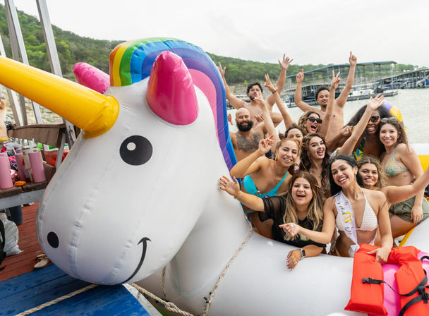 Cheerful group of friends on a giant inflatable unicorn float at a lakeside dock party, posing in swimsuits and cheering