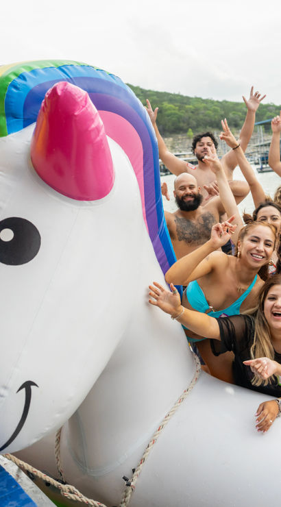 Cheerful group of friends on a giant inflatable unicorn float at a lakeside dock party, posing in swimsuits and cheering