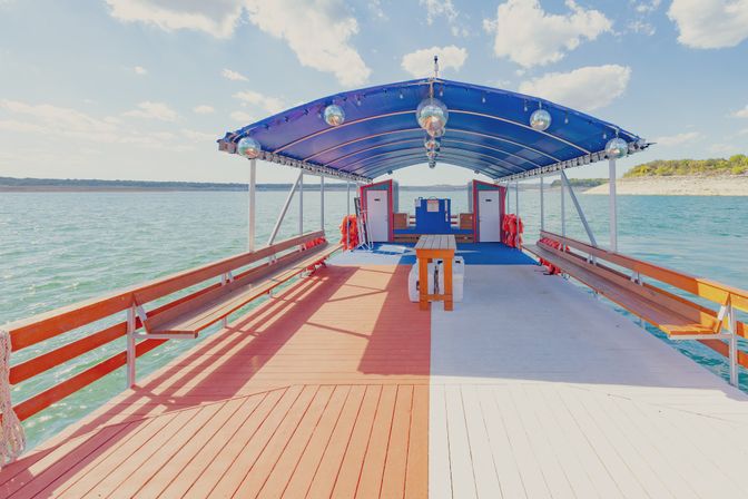 Bright party boat deck on a calm lake with a blue canopy and disco balls, long wooden benches, small picnic table and sunny shoreline in the distance.