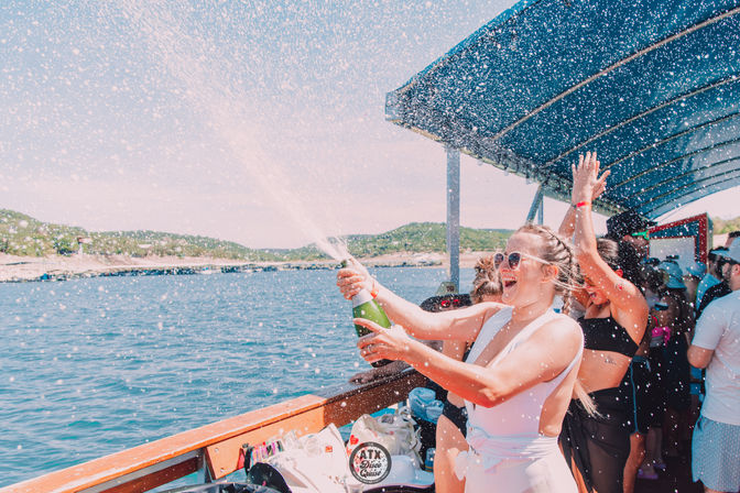 Sunny daytime boat party with passengers in swimsuits spraying champagne on the deck, water droplets flying and a hilly shoreline in the background.