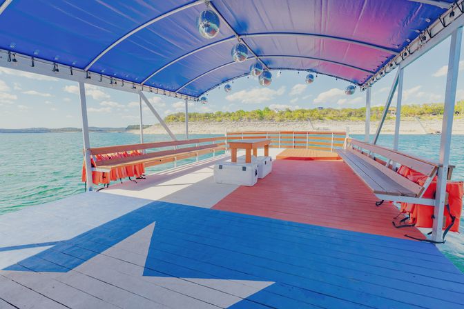 Covered lake party pontoon with benches, orange life jackets, hanging disco balls and a red-white-blue deck featuring a large star, overlooking a tree-lined shoreline on a sunny day.