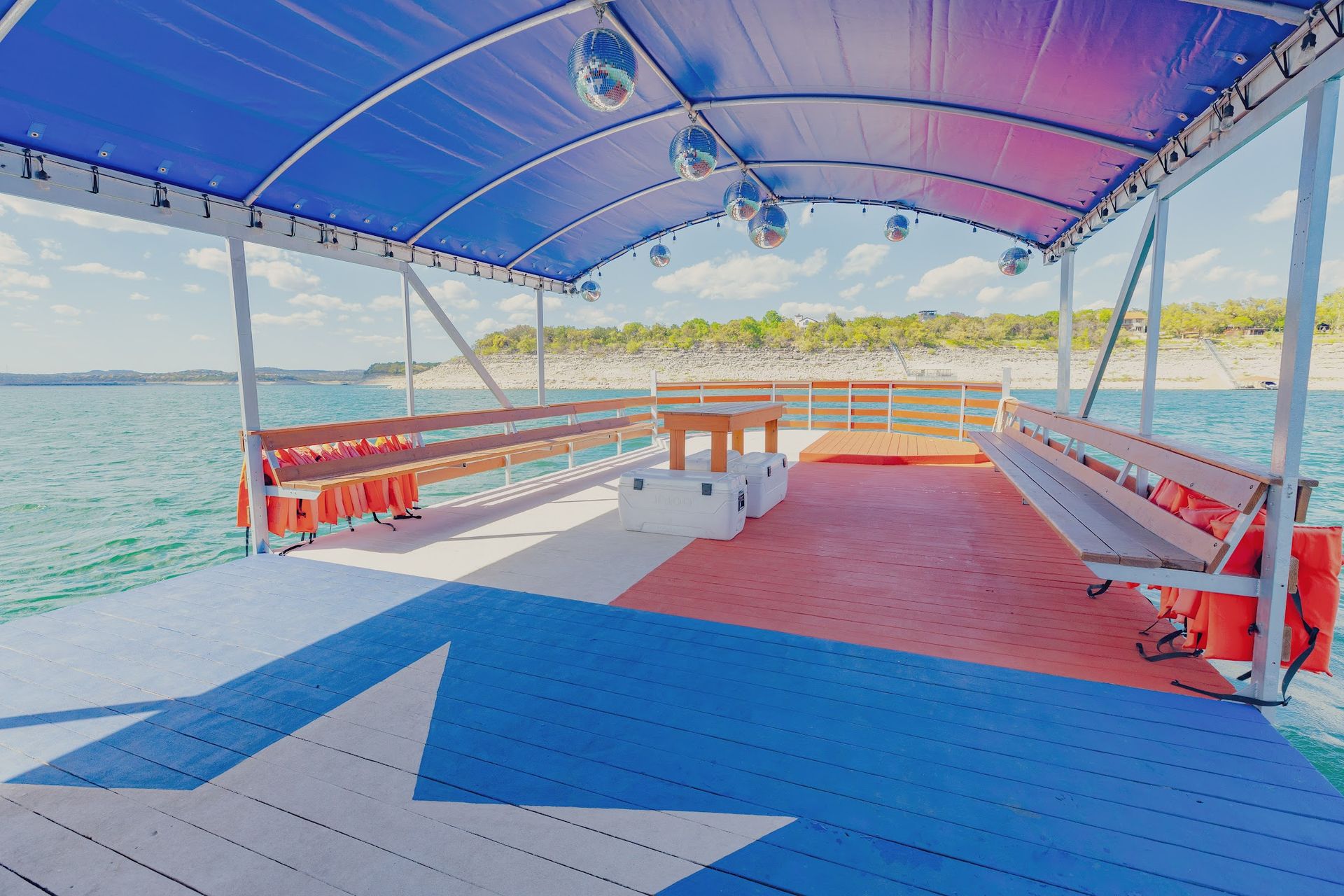 Covered lake party pontoon with benches, orange life jackets, hanging disco balls and a red-white-blue deck featuring a large star, overlooking a tree-lined shoreline on a sunny day.