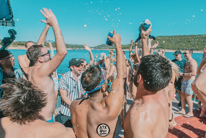Sunny summer boat party on a lake with a crowd of people cheering, raising drinks and splashing water against turquoise water and green shoreline