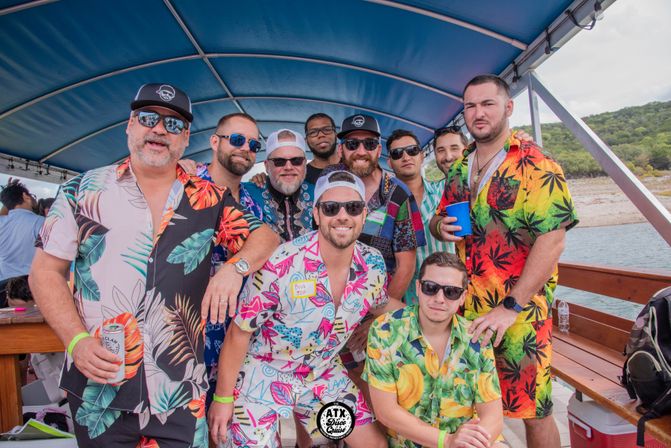 Group of men in colorful Hawaiian shirts and sunglasses smiling and posing on a covered party boat over a lake with a tree-lined shoreline in the background