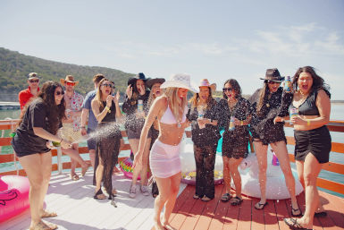 Energetic lake party on a wooden dock — a group of friends in swimsuits and cowboy hats laughing as champagne sprays over colorful inflatable floats, sunlit water and green hills in the background.