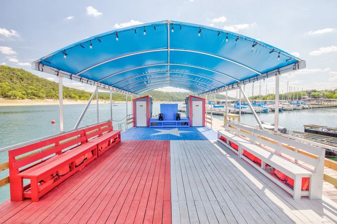 Patriotic-themed covered lake dock and marina with red, white, and blue wood deck featuring a large star, benches lining both sides, a blue canopy overhead and sailboats and tree-lined shore in the sunny background.