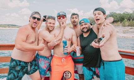 Six men in colorful swim trunks cheer and pose with canned drinks on a sunny summer boat over a tree-lined lake.