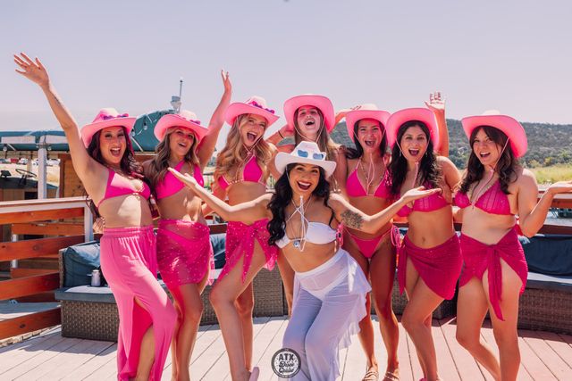 Eight women in pink bikinis and cowgirl hats cheer and pose on a sunny lakeside deck for a playful bachelorette-style celebration, bright summer party vibes.