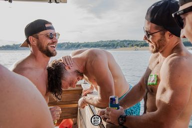 Shirtless friends laughing on a sunny lake boat ride, two in backward caps and sunglasses, one leaning over the rail