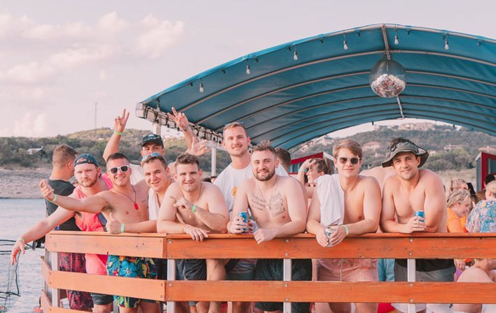 Group of young men at a sunny lake party on a covered pontoon boat, shirtless and smiling while leaning on a wooden railing and holding canned drinks under a blue canopy