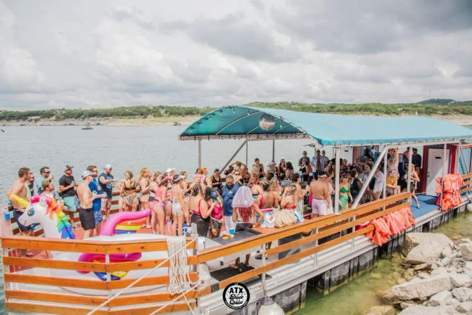 Crowded lake party on a covered pontoon boat with dozensof people in swimsuits, colorful inflatable unicorn float, turquoise canopy, life jackets on railing and rocky shoreline in the background