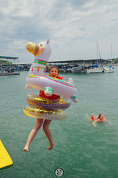 Person jumping off a dock into a lake marina wearing a life jacket and stacked inflatables — a colorful llama float with gold-glitter rings, sailboats and covered slips in the background