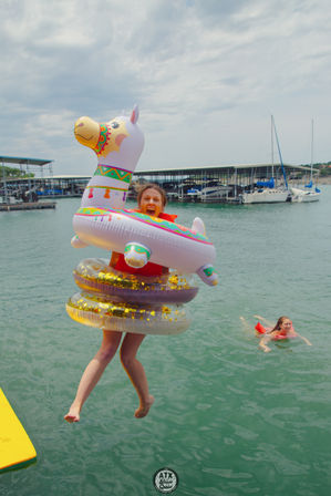 Person jumping off a dock into a lake marina wearing a life jacket and stacked inflatables — a colorful llama float with gold-glitter rings, sailboats and covered slips in the background