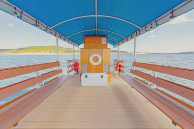 Covered wooden boat deck with long benches under a bright blue canopy, white lifebuoy and cooler centered, open view over calm lake water toward a green shoreline on a sunny day