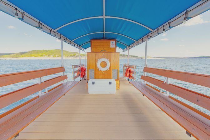 Covered wooden boat deck with long benches under a bright blue canopy, white lifebuoy and cooler centered, open view over calm lake water toward a green shoreline on a sunny day