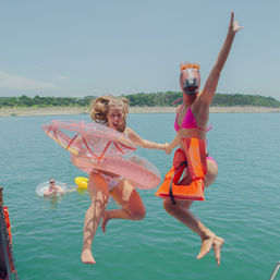Two people mid-jump off a boat into a sunny lake — one in a pink bikini wearing a horse mask and orange life jacket, the other clutching pink inflatable rings with a shoreline and a floater in the water behind them.