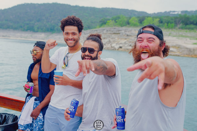 Four friends laughing and pointing at the camera on a sunny lake boat near a rocky shoreline, holding cold drinks for a summer boat party.