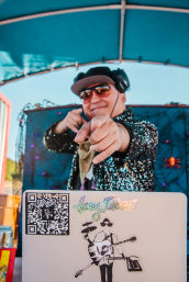 Outdoor daytime DJ in sunglasses and headphones pointing at the camera from a decorated booth under a blue canopy, wearing a sparkly sequined jacket with a laptop showing music stickers and a QR code visible.