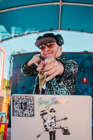 Outdoor daytime DJ in sunglasses and headphones pointing at the camera from a decorated booth under a blue canopy, wearing a sparkly sequined jacket with a laptop showing music stickers and a QR code visible.