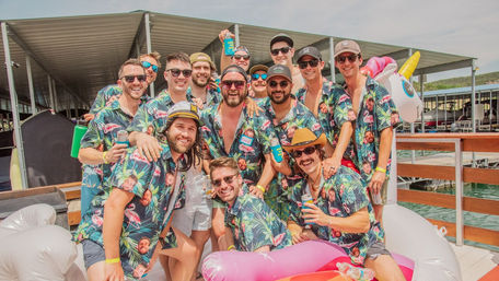 Group of men in matching tropical shirts smiling with drinks on a sunny lake dock at a marina, surrounded by colorful inflatable floats and a unicorn tube