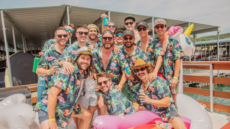 Group of men in matching tropical shirts smiling with drinks on a sunny lake dock at a marina, surrounded by colorful inflatable floats and a unicorn tube