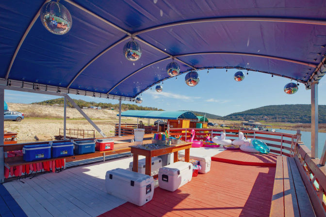 Covered lakeside dock with blue canopy and mirrored disco balls, coolers and life jackets along the rail, a table with drinks and colorful inflatable swan and donut floats, overlooking a calm lake and rolling hills.