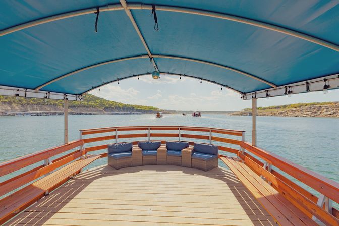 Covered wooden boat deck with blue canopy, wicker lounge chairs and bench seating facing a sunny lake view with rocky shoreline and distant boats