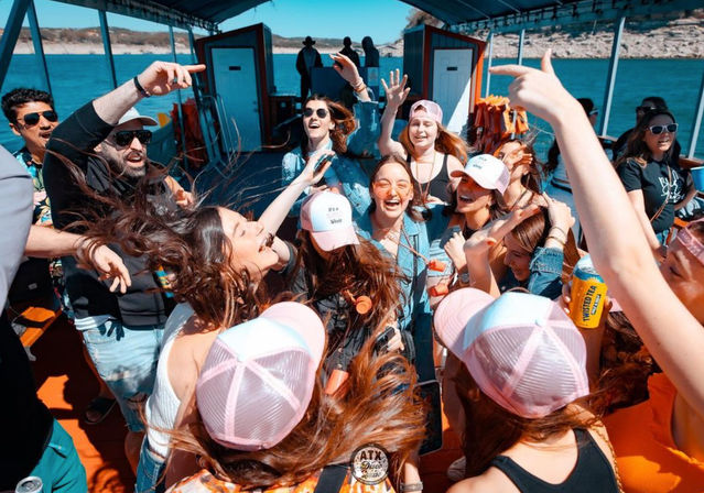 Group of friends dancing and laughing on a crowded party boat at a sunny lake, many wearing pink trucker hats and holding drinks.