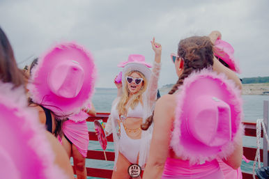 Women at a lively summer boat party near the shoreline, wearing pink feathered cowboy hats and matching outfits; central woman in a white swimsuit and heart-shaped sunglasses cheering with her arm raised.