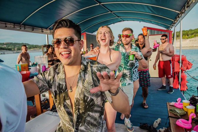 Friends partying on a covered pontoon boat at a sunny lake — a conga line of young adults in tropical shirts and swimsuits holding colorful cups and inflatable flamingos.