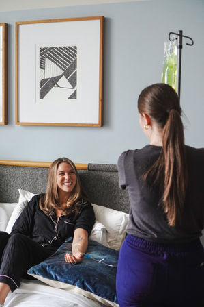 Smiling woman on a bed receiving an at‑home IV infusion as a caregiver adjusts the IV pole in a bright bedroom with framed abstract art.
