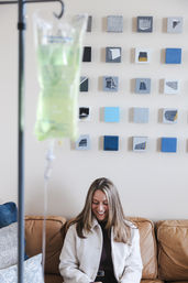 Smiling woman seated on a tan leather couch in a cozy clinic lounge, IV infusion bag in the foreground and a grid of blue-gray square wall art behind.