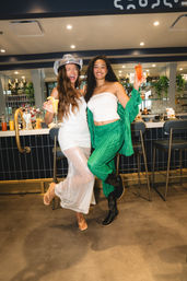 Two friends laughing and raising colorful cocktails at a modern bar — one in a sparkly white dress and silver cowboy hat, the other in a green outfit and boots, celebrating a night out.