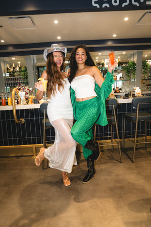 Two friends laughing and raising colorful cocktails at a modern bar — one in a sparkly white dress and silver cowboy hat, the other in a green outfit and boots, celebrating a night out.