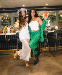 Two friends laughing and posing with colorful cocktails—one in a sheer white dress and cowboy hat, the other in green pants and a white tube top—lifting a leg in front of a modern indoor cocktail bar with stools and a backlit bottle display.