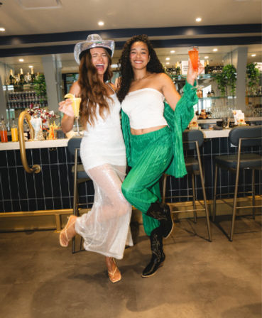 Two friends laughing and posing with colorful cocktails—one in a sheer white dress and cowboy hat, the other in green pants and a white tube top—lifting a leg in front of a modern indoor cocktail bar with stools and a backlit bottle display.
