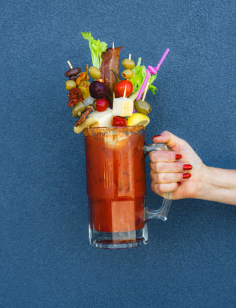 Hand holding oversized Bloody Mary mug with seasoned rim, loaded with celery stalk, crispy bacon, skewers of olives, cherry tomatoes, cheese and pickles, and a pink straw against a blue wall