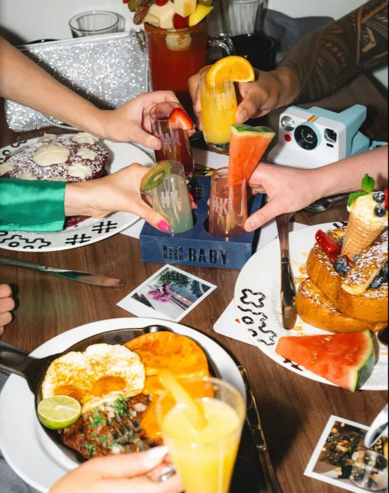 Several hands clinking colorful brunch cocktails and mimosas over a wooden table with a skillet of eggs and steak, stacked French toast with berries and an ice-cream cone, watermelon slices, fruit-garnished shot glasses and a Polaroid camera — lively weekend brunch scene.