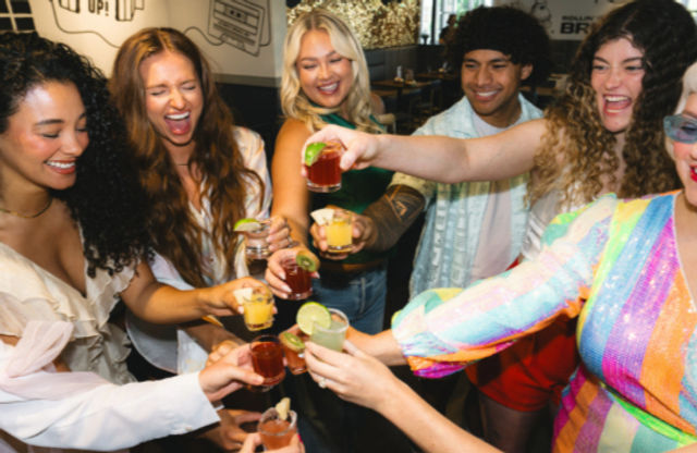 Smiling friends toasting colorful cocktails and shot glasses in a lively indoor bar celebration.