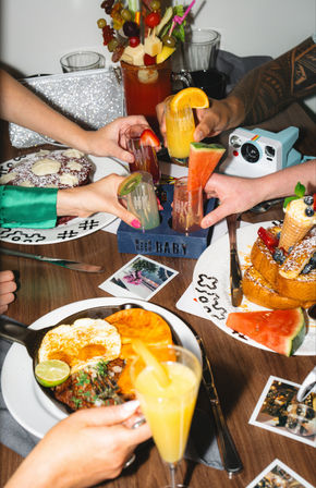 Brunch table with friends toasting colorful cocktails garnished with watermelon, kiwi and orange, surrounded by pancakes with cream, stacked French toast with berries and a skillet breakfast with fried egg and lime